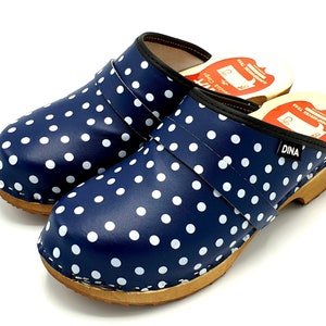 Könnte beinhalten: Ein Paar blaue Clogs mit weißen Punkten und einer Holzsohle. Die Clogs haben einen schwarzen Riemen über dem Oberteil und der Text "DINA" ist seitlich aufgedruckt.