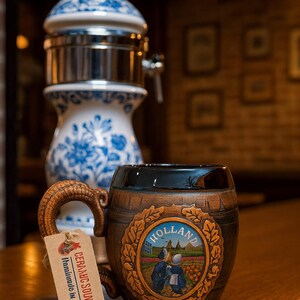 Op de afbeelding: Een keramische souvenirbeker met een vatontwerp en een handvat. De beker heeft een geschilderde scène met het woord "HOLLAND" en een label met de tekst "CERAMIC SOUVENIRS Handmade in Europe". Op de achtergrond staat een blauw-witte keramische dispenser.