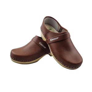 Puede incluir: Un par de zuecos de cuero marrón con una correa y el nombre de la marca "Simson" impreso en blanco. Los zuecos tienen una suela de color claro con costuras blancas. Los zapatos están diseñados para la comodidad y el soporte.