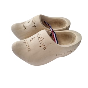 Könnte beinhalten: Zwei hellbraune Holzschuhe mit eingravierten Namen und einem Datum. Die Schuhe haben einen ausgehöhlten Innenraum und ein rot-weiß-blaues Band. Auf einem Schuh ist das Datum '11-10-2024' zu sehen.