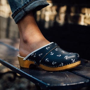 Puede incluir: Un par de zuecos azules y blancos con estampado de ancla. Los zuecos tienen una base de madera y una parte superior de cuero. Los zuecos son usados por una persona que lleva pantalones vaqueros azules.