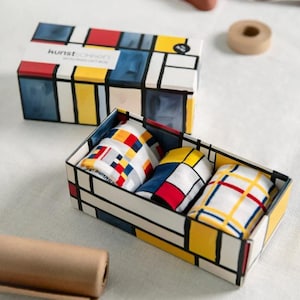 Op de afbeelding: Een geschenkdoosset met een Mondriaan-stijl ontwerp. De doos is wit met zwarte lijnen en kleurrijke blokken rood, blauw en geel. Binnenin zitten drie items met patronen. Op het deksel staat "kunstkonzept" en "MEMORIAL GIFT BOX."