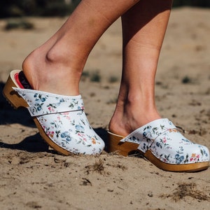 Puede incluir: Un par de zuecos blancos con estampado floral. Los zuecos tienen una base de madera y una correa en la parte superior.