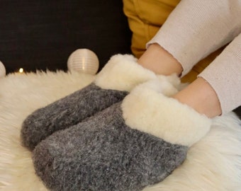 May include: Grey wool slippers with a white sheepskin lining. The slippers are being worn by a person sitting on a white fluffy rug.