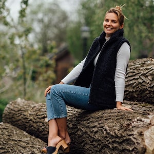 Op de afbeelding: Een zwart sherpa fleece vest gedragen over een wit shirt met lange mouwen en ribbelstructuur, gecombineerd met een blauwe spijkerbroek en zwarte leren klompen met houten zolen. Het vest heeft een hoge kraag, een rits en twee zijzakken.
