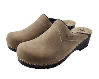 May include: A pair of tan suede clogs with black soles. The clogs have a rounded toe and a low heel. The word "DINA" is stitched on the side of each shoe. The clogs are a slip-on style.