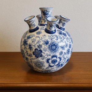 Puede incluir: Un jarrón redondo de cerámica blanca con un diseño floral azul. El jarrón tiene múltiples aberturas para flores, un cuerpo bulboso y un borde azul oscuro. El diseño presenta flores y follaje azules detallados.