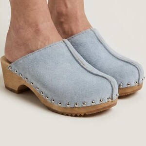 May include: Light blue suede clogs with wooden soles. The clogs have a rounded toe and are adorned with silver-colored studs around the sole. The wooden heel is approximately 2 inches high.
