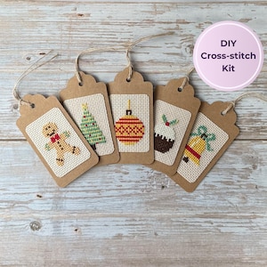 May include: Five brown cardboard gift tags featuring cross-stitch designs of a gingerbread person, a Christmas tree, a striped ornament, a pudding, and a bell. Each tag has a jute string loop at the top. Text reads: DIY Cross-stitch Kit.
