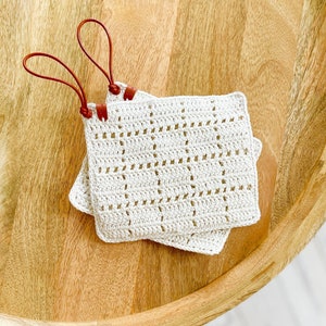 May include: Two white crocheted pot holders with brown leather loops. The pot holders are on a wooden surface.