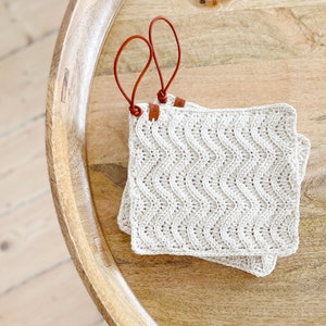 May include: Two crocheted pot holders with a wavy pattern and brown leather loops. The pot holders are on a wooden surface.