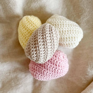 Op de afbeelding: Vier handgemaakte gehaakte eieren in pastelkleuren: geel, beige, wit en roze. De eieren zijn gerangschikt op een neutraal gekleurde stof. Elk ei heeft een gestructureerd, geribbeld ontwerp dat het gedetailleerde haakwerk laat zien.