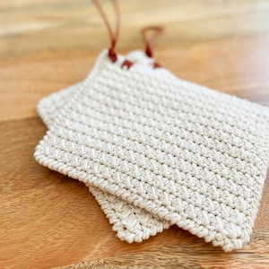 Peut inclure: Deux maniques en crochet blanc cassé avec un motif texturé sont empilées sur une surface en bois. Chaque manique a un bord festonné et une boucle en cuir marron pour suspendre. Les maniques sont conçues pour la cuisine.