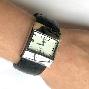 May include: A silver-toned rectangular watch with a white face and black leather band. The watch face displays the brand name "Zella Park" and the word "QUARTZ". The black band has a crocodile skin texture.