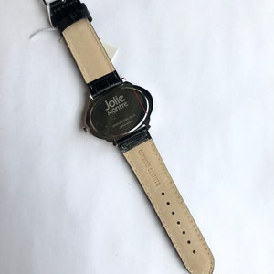 May include: Black leather watch with a stainless steel back. The watch band is black with a crocodile texture. The watch face is oval-shaped and has the words "Jolie Montre" printed on it. The watch band is genuine leather.