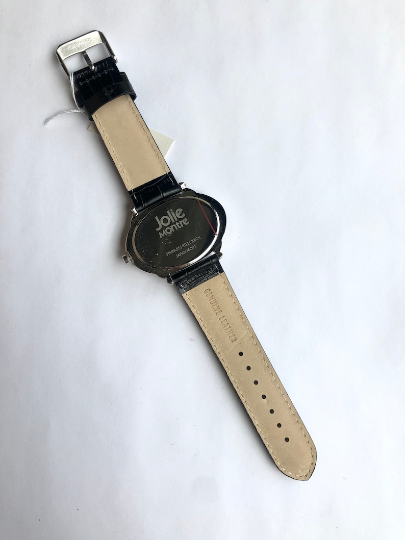 May include: Black leather watch with a stainless steel back. The watch band is black with a crocodile texture. The watch face is oval-shaped and has the words "Jolie Montre" printed on it. The watch band is genuine leather.
