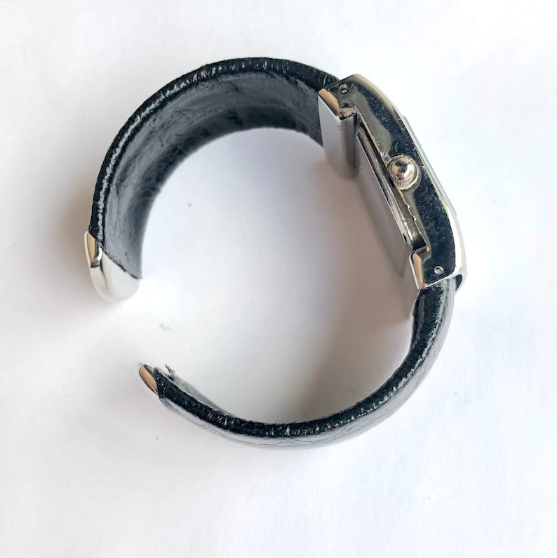 May include: A black leather cuff watch with a rectangular silver-toned watch face. The watch band is a wide, curved cuff with silver metal end caps. The watch is set against a white background.