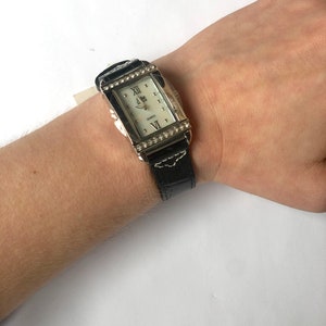 Puede incluir: Un reloj de cuarzo rectangular con caja plateada y esfera blanca. El reloj tiene una correa de cuero negro con costuras blancas y est&aacute; adornado con peque&ntilde;os cristales transparentes alrededor de la esfera. El reloj est&aacute; en una mu&ntilde;eca.