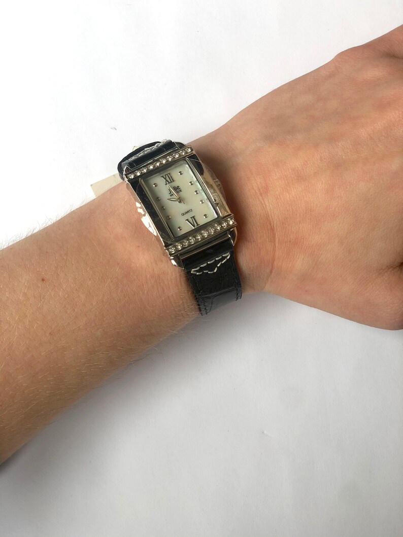 Puede incluir: Un reloj de cuarzo rectangular con caja plateada y esfera blanca. El reloj tiene una correa de cuero negro con costuras blancas y est&aacute; adornado con peque&ntilde;os cristales transparentes alrededor de la esfera. El reloj est&aacute; en una mu&ntilde;eca.