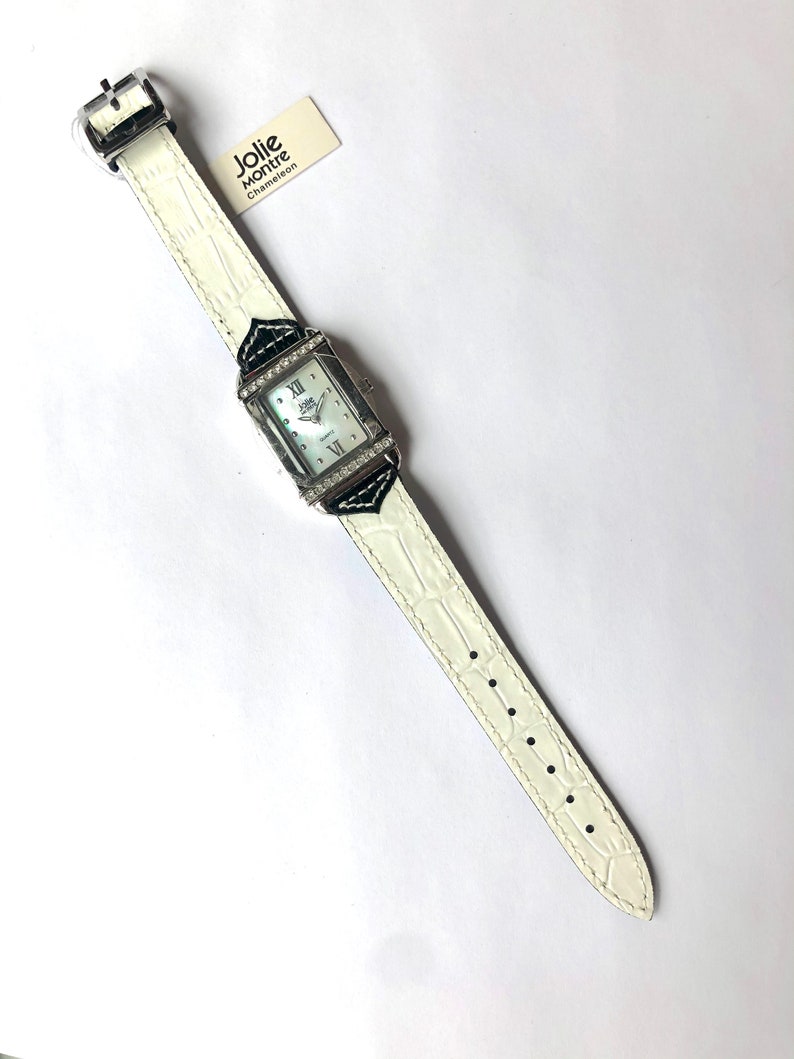 Puede incluir: Un reloj blanco con una esfera rectangular y una correa blanca. La esfera del reloj tiene n&uacute;meros romanos y est&aacute; rodeada de peque&ntilde;os cristales. La correa tiene un patr&oacute;n de cocodrilo. El reloj tiene una etiqueta que dice "Jolie Montre Chameleon".