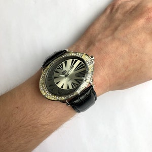 May include: Oval-shaped wristwatch with a silver face and black leather band. The watch has a silver bezel encrusted with clear crystals. The watch face features black radial lines and the brand name "Jolie."