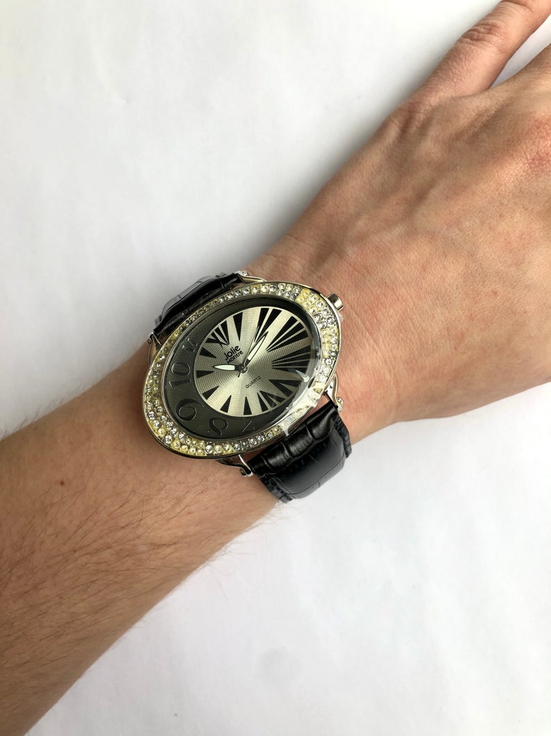 May include: Oval-shaped wristwatch with a silver face and black leather band. The watch has a silver bezel encrusted with clear crystals. The watch face features black radial lines and the brand name "Jolie."