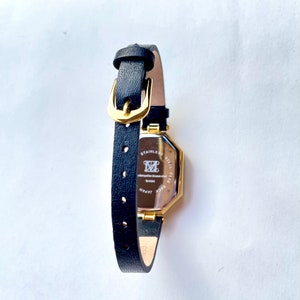 May include: A black leather watch with a gold-colored rectangular face. The watch has a black leather band with a gold buckle. The back of the watch has text that reads "STAINLESS STEEL CASE" and "JAPAN".