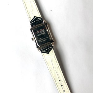 Puede incluir: Un reloj Jolie Montre Chameleon rectangular con una correa de cuero blanco texturizado. La esfera del reloj es plateada con detalles en negro. La parte trasera del reloj es de acero inoxidable y el movimiento es japon&eacute;s. Una etiqueta con el nombre de la marca est&aacute; adjunta.