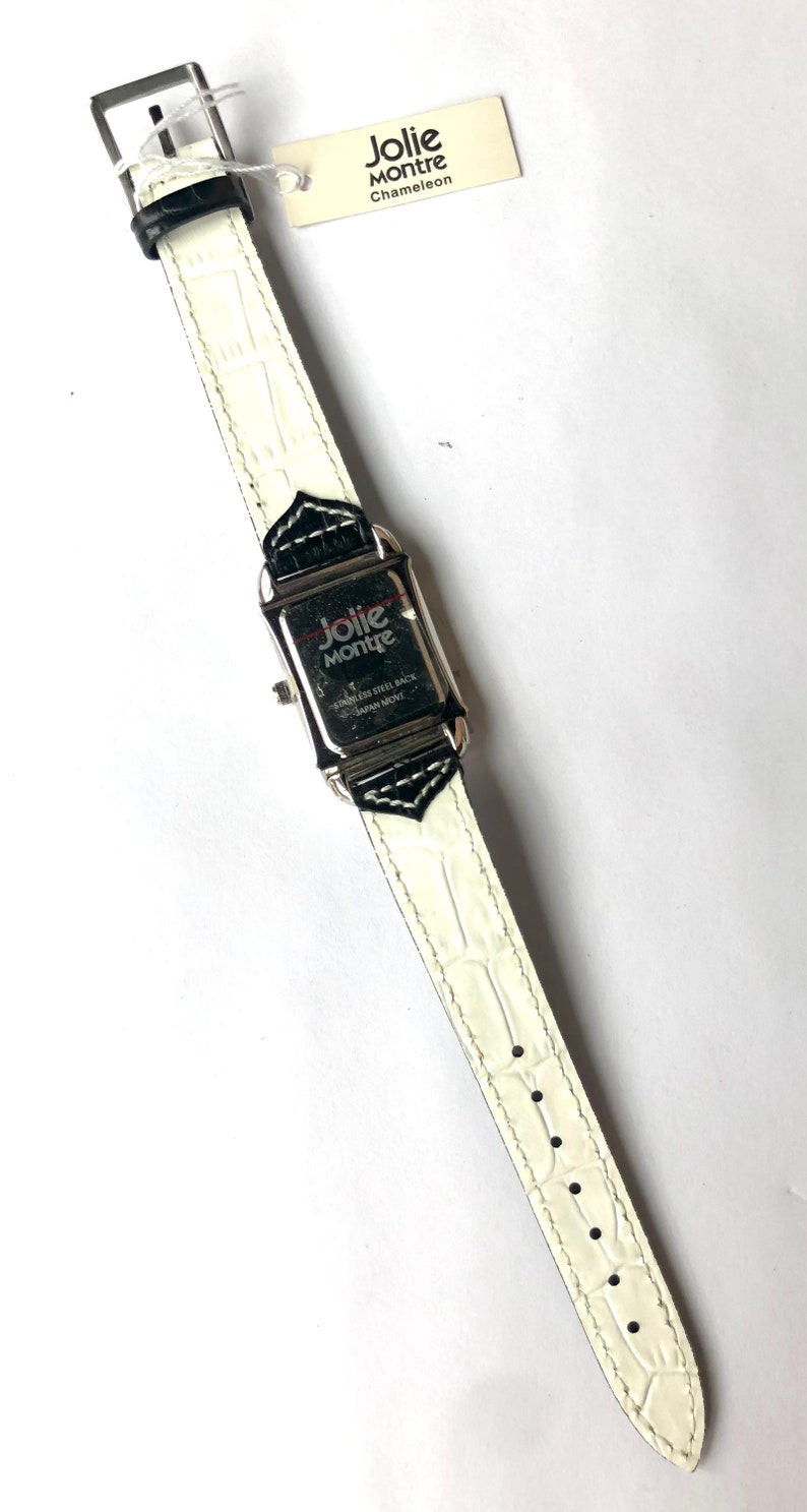 Puede incluir: Un reloj Jolie Montre Chameleon rectangular con una correa de cuero blanco texturizado. La esfera del reloj es plateada con detalles en negro. La parte trasera del reloj es de acero inoxidable y el movimiento es japon&eacute;s. Una etiqueta con el nombre de la marca est&aacute; adjunta.