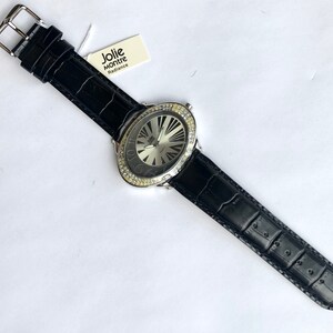May include: A black leather watch with a silver face and a bezel encrusted with clear crystals. The watch has a black leather band with a buckle closure. The watch face has a radial design. The tag reads "Jolie Montre Radiance."
