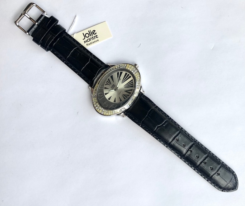 May include: A black leather watch with a silver face and a bezel encrusted with clear crystals. The watch has a black leather band with a buckle closure. The watch face has a radial design. The tag reads "Jolie Montre Radiance."