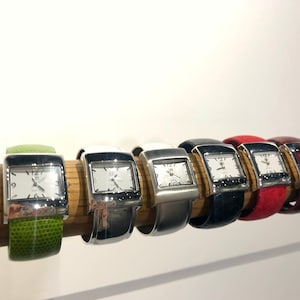 May include: A collection of rectangular-faced watches with silver-tone cases and white dials, each displaying the time. The watches have various colored bands, including green, white, silver, black, red, and burgundy. The watches are displayed on a wooden rod.