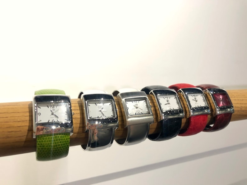 May include: A collection of rectangular-faced watches with silver-tone cases and white dials, each displaying the time. The watches have various colored bands, including green, white, silver, black, red, and burgundy. The watches are displayed on a wooden rod.