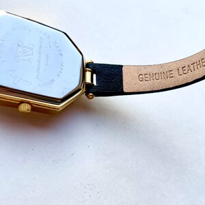 May include: Close-up of a gold-toned watch with a black leather band. The watch face is octagonal. The back of the watch reads "Stainless Steel Case Back Japan Movt Quartz". The band is stamped "Genuine Leather".