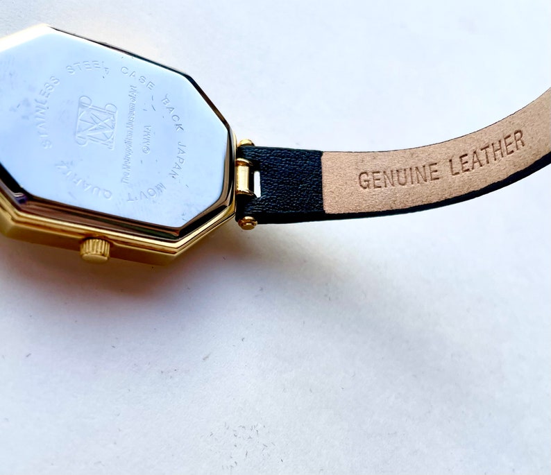 May include: Close-up of a gold-toned watch with a black leather band. The watch face is octagonal. The back of the watch reads "Stainless Steel Case Back Japan Movt Quartz". The band is stamped "Genuine Leather".