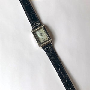 Puede incluir: Un reloj Jolie Montre Chameleon de cuarzo rectangular con una correa de cuero negro con estampado de cocodrilo. La esfera del reloj es plateada con n&uacute;meros romanos y peque&ntilde;os cristales alrededor del bisel. Una etiqueta con el nombre de la marca est&aacute; adjunta.