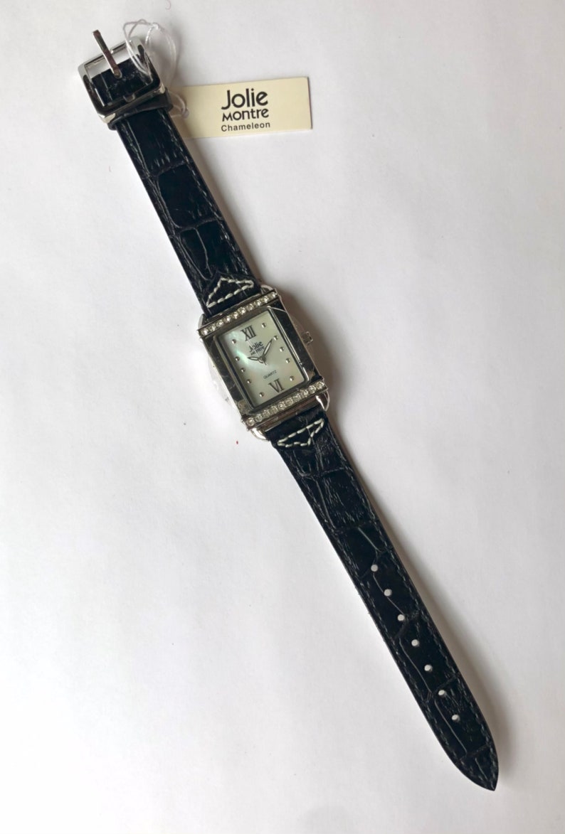 Puede incluir: Un reloj Jolie Montre Chameleon de cuarzo rectangular con una correa de cuero negro con estampado de cocodrilo. La esfera del reloj es plateada con n&uacute;meros romanos y peque&ntilde;os cristales alrededor del bisel. Una etiqueta con el nombre de la marca est&aacute; adjunta.