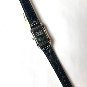 Puede incluir: Un reloj de cuero negro con una esfera rectangular plateada. La correa del reloj tiene una textura de cocodrilo. La esfera del reloj tiene las palabras "Jolie Montre" y "Stainless Steel Back Japan Movt". Una etiqueta blanca est&aacute; adherida a la correa.