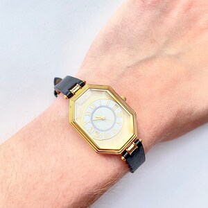 May include: A gold-toned watch with a unique octagonal face and a black leather band. The watch face features Roman numerals and gold hands. The watch is approximately 1 inch in diameter.