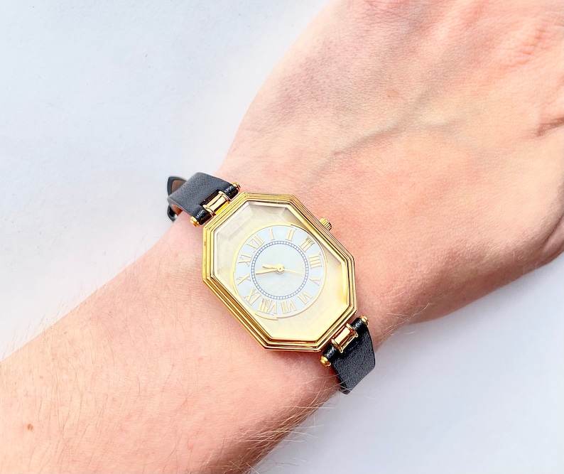 May include: A gold-toned watch with a unique octagonal face and a black leather band. The watch face features Roman numerals and gold hands. The watch is approximately 1 inch in diameter.