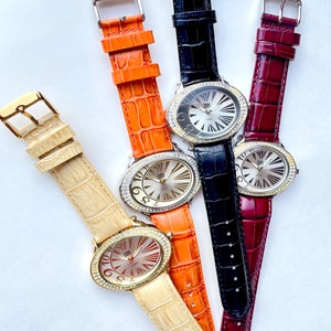 May include: Four oval-faced watches with leather bands in cream, orange, black, and burgundy. Each watch has a silver dial with gold-tone hands and hour markers. The watch faces are surrounded by a ring of clear crystals.