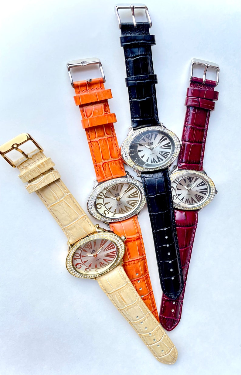 May include: Four oval-faced watches with leather bands in cream, orange, black, and burgundy. Each watch has a silver dial with gold-tone hands and hour markers. The watch faces are surrounded by a ring of clear crystals.