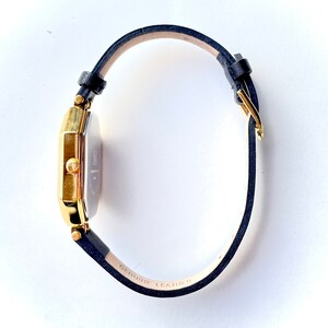 May include: A gold-toned rectangular watch with a navy blue leather band. The watch face is silver with black hands and markers. The band is labeled "GENUINE LEATHER". The watch is a classic accessory.