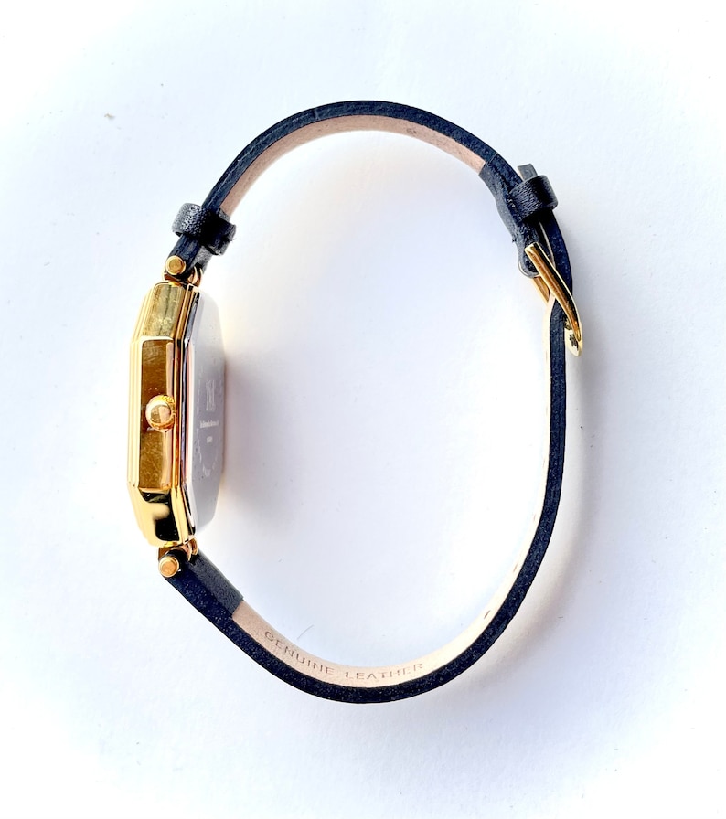 May include: A gold-toned rectangular watch with a navy blue leather band. The watch face is silver with black hands and markers. The band is labeled "GENUINE LEATHER". The watch is a classic accessory.