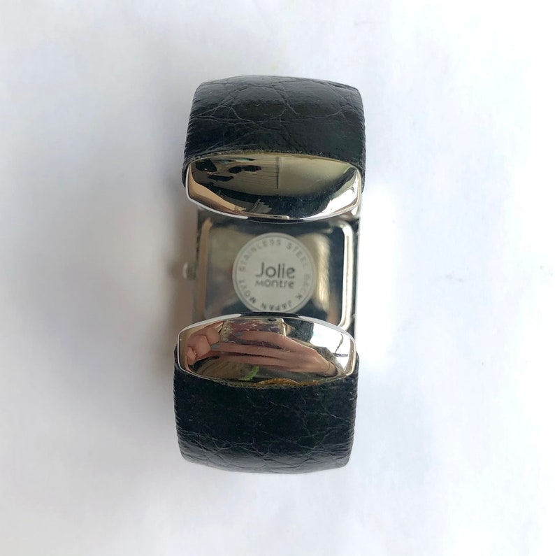 May include: A black leather cuff watch with a silver-tone stainless steel case. The watch face is visible, with the brand name "Jolie Montre" and the words "Stainless Steel Back Japan Movement" printed on it. The watch is on a white surface.