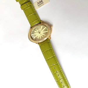 May include: A gold-toned Jolie Montre watch with a light green leather band. The watch face is oval-shaped, surrounded by crystals, and features large numerals. The band has a crocodile texture. The tag reads "Jolie Montre Radiance."