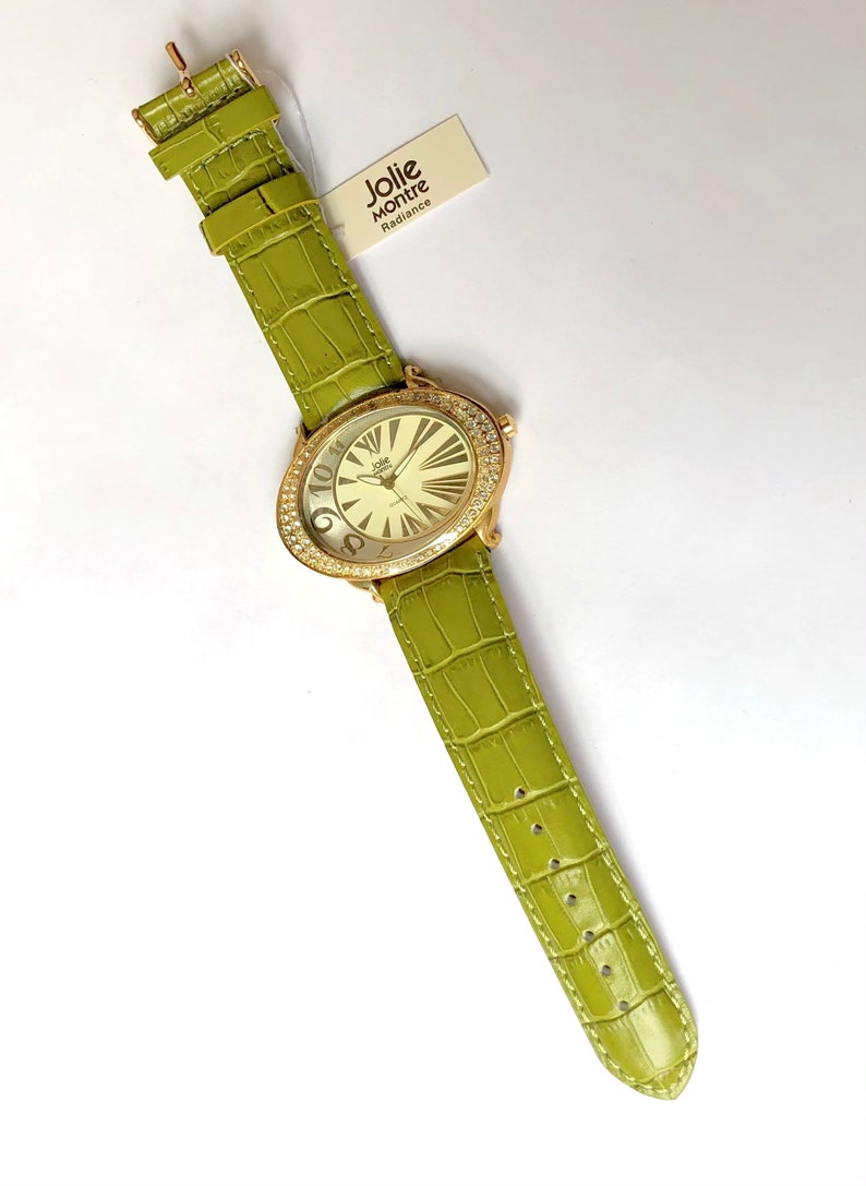 May include: A gold-toned Jolie Montre watch with a light green leather band. The watch face is oval-shaped, surrounded by crystals, and features large numerals. The band has a crocodile texture. The tag reads "Jolie Montre Radiance."