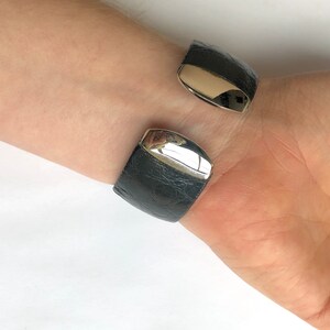 May include: A black leather cuff bracelet with silver-colored metal accents. The bracelet has a textured surface and a hinged design. The silver metal is polished and reflects light. The bracelet is worn on a wrist.