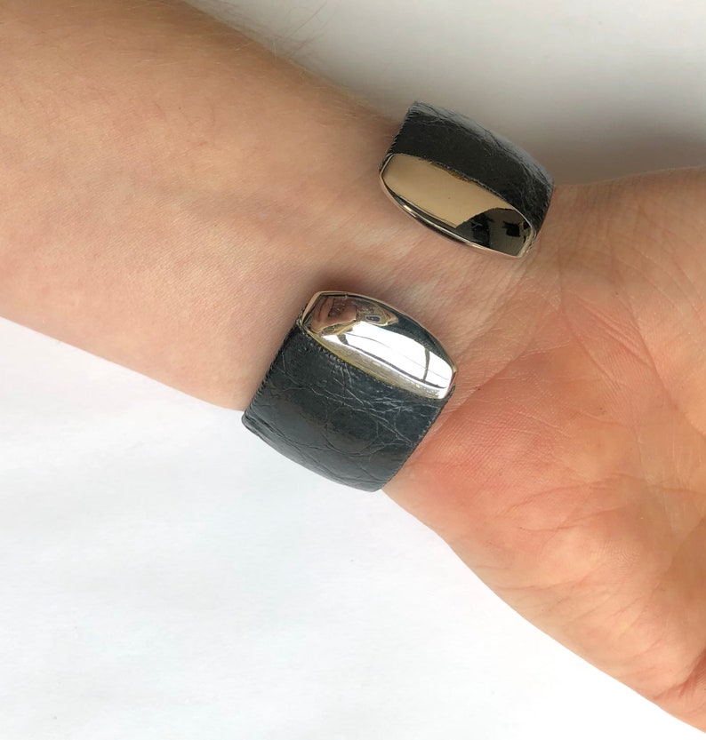 May include: A black leather cuff bracelet with silver-colored metal accents. The bracelet has a textured surface and a hinged design. The silver metal is polished and reflects light. The bracelet is worn on a wrist.