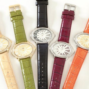 May include: Five oval-faced watches with rhinestone bezels and textured leather straps in beige, green, black, burgundy, and orange. The watch faces have a sunburst design and the straps have gold or silver buckles.